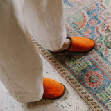 Person in cream trousers wearing orange lambskin slippers on a patterned rug
