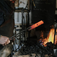 A man standing at a fire forge with a red hot piece of steel in tongs. 