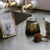Bag of Prima coffee, stovetop espresso maker and a coffeee scoop on stainless steel benchtop. 